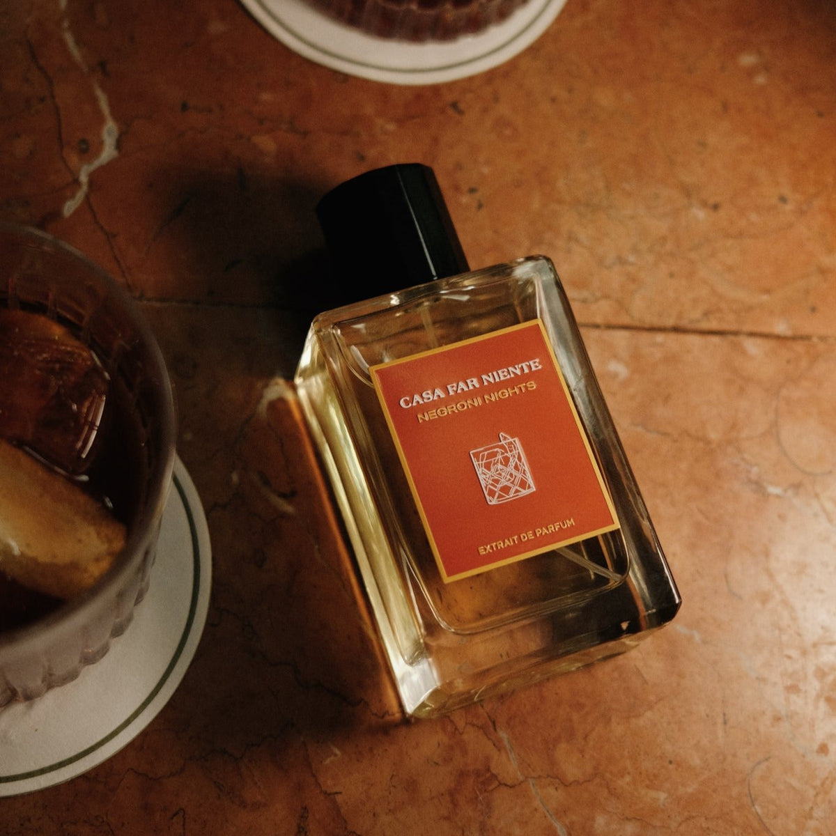 Casa Far Niente Negroni Nights Flakon mit roter Plakette, Lifestyle-Aufnahme auf rotem Steintisch liegend mit Cocktailglass